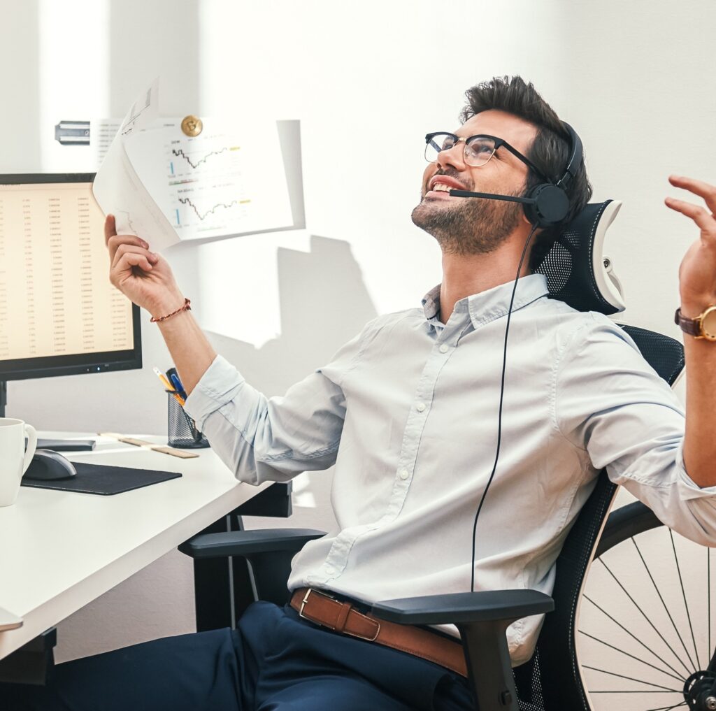 Great! Happy And Successful Bearded Trader In Headset Holding Financial Report, Talking With Client And Gesturing While Sitting In Front Of Monitor Screens In The Office.