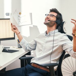 Great! Happy And Successful Bearded Trader In Headset Holding Financial Report, Talking With Client And Gesturing While Sitting In Front Of Monitor Screens In The Office.