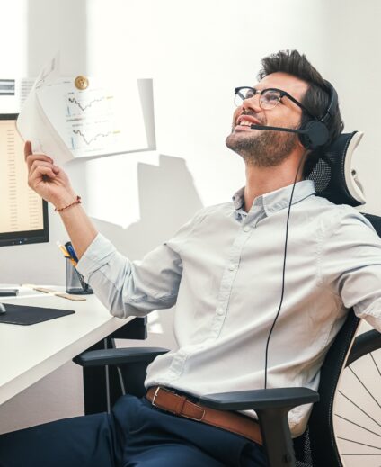 Great! Happy And Successful Bearded Trader In Headset Holding Financial Report, Talking With Client And Gesturing While Sitting In Front Of Monitor Screens In The Office.