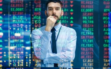 Stock Market Top Trader Looks At Projected Ticker Numbers And Graphs Running, Analysing Data To Make Best Sell. Behind Him Room Full Of Screens And Statistics.