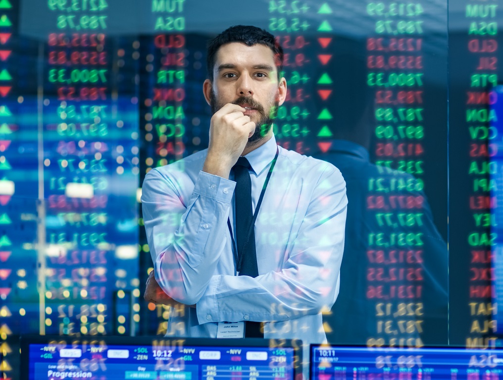 Stock Market Top Trader Looks At Projected Ticker Numbers And Graphs Running, Analysing Data To Make Best Sell. Behind Him Room Full Of Screens And Statistics.